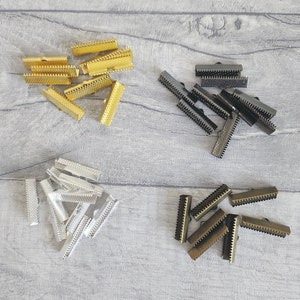 May include: Assortment of metal slider buckles in four colours: gold, silver, black, and bronze. The buckles are arranged in four groups on a wooden surface.
