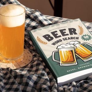 Könnte beinhalten: Ein hohes Bierglas mit Schaum steht auf einem Untersetzer neben einem Buch mit dem Titel "Beer Word Search". Das Buch zeigt Illustrationen von Bierkrügen und den Text "Beers, Breweries & More."
