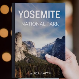 May include: A book titled "YOSEMITE NATIONAL PARK WORD SEARCH" held in a person's hand. The cover displays a scenic landscape of mountains, trees, and a blue sky. The book has a black spine and a glossy cover.