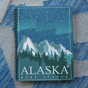 May include: Spiral-bound notebook with an Alaska-themed cover. The cover features a landscape illustration of mountains, trees, and a starry sky in shades of blue and green. The word "ALASKA" is printed in white, with "WORD SEARCH" below.
