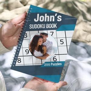 May include: A blue and gray spiral bound book with the title "John's Sudoku Book" and a photo of a couple on the cover. The book contains 200 puzzles.