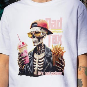 Puede incluir: Camiseta blanca con un diseño de esqueleto que lleva una chaqueta de cuero, gafas de sol y una gorra roja. El esqueleto sostiene un batido y patatas fritas, con el texto "Dad Tax" y "It's not poison."