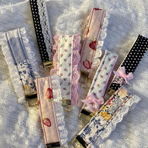 May include: A collection of fabric wrist keychains in various patterns. Designs include strawberries, floral prints, and polka dots, trimmed with lace. Each keychain has a gold-colored metal ring for attaching keys.