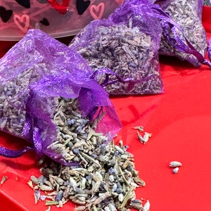 Puede incluir: Tres bolsas de malla púrpura llenas de flores de lavanda secas. Las bolsas están sobre una superficie roja.