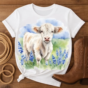 Op de afbeelding: Wit T-shirt met een aquarel illustratie van een witte koe in een veld met blauwe bloemen. De koe heeft lichtbruine oren en een roze neus. De achtergrond omvat een blauwe lucht en groen gras.