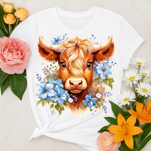 Op de afbeelding: Wit T-shirt met een aquarel illustratie van een Schotse Hooglander. De koe heeft bruine vacht, grote hoorns en is omringd door blauwe bloemen en bladeren. Het ontwerp heeft een rustieke en charmante uitstraling.