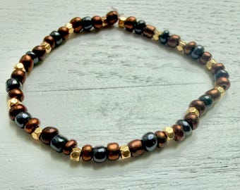 May include: A beaded bracelet featuring a repeating pattern of metallic copper-colored round beads, dark charcoal grey round beads, and small, faceted gold-toned spacer beads arranged in a circular loop on a light-colored surface.