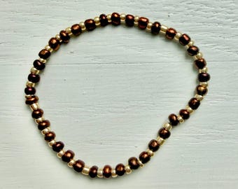 May include: A circular beaded bracelet featuring an alternating pattern of small, metallic bronze-colored beads and smaller, smooth gold-toned beads arranged in a continuous loop on a flexible cord.