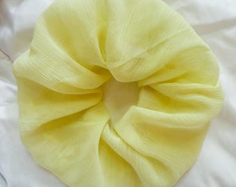 May include: A pale yellow hair scrunchie made of a lightweight, semi-sheer fabric with a fine, crinkled texture. The circular accessory features gathered folds throughout, creating a voluminous, ruffled appearance around the central opening.