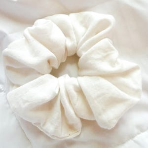 May include: A solid off-white fabric hair scrunchie with a gathered, ruffled texture. The accessory features a circular shape with a soft, matte finish and a consistent, neutral light cream color throughout the entire piece of hair jewelry.