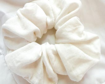 May include: A solid off-white fabric hair scrunchie with a gathered, ruffled texture. The accessory features a circular shape with a soft, matte finish and a consistent, neutral light cream color throughout the entire piece of hair jewelry.