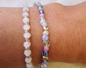 May include: Two beaded bracelets. One bracelet features uniform white faceted beads interspersed with small metallic gold spacers. The second bracelet consists of a multi-colored pattern of pink, light blue, dark blue, and gold beads in various shapes.