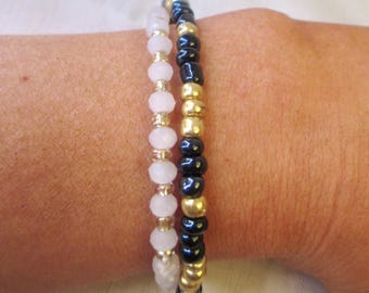 May include: Two beaded bracelets worn together. One bracelet features alternating pale pink faceted beads and small gold-tone spacers. The second bracelet consists of alternating glossy black round beads and metallic gold-tone beads in a repeating pattern.