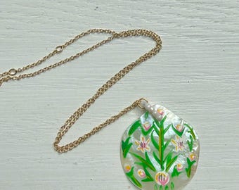 May include: A gold-toned metal chain necklace featuring a round, iridescent mother-of-pearl pendant. The pendant is hand-painted with a vibrant green, pink, and white floral design, suspended from the chain by a small, white, cylindrical bead.