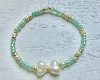 May include: A beaded bracelet featuring a repeating pattern of light mint green glass beads, small metallic gold spacer beads, and white spherical beads. The center of the bracelet showcases two white heart-shaped beads separated by a gold bead.