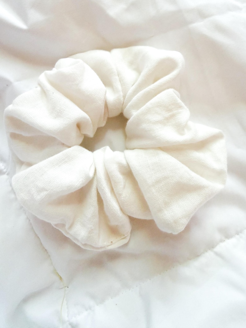 May include: A solid off-white fabric hair scrunchie with a gathered, ruffled texture. The accessory features a circular shape with a soft, matte finish and a consistent, neutral light cream color throughout the entire piece of hair jewelry.