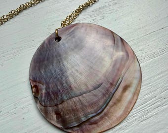 May include: A circular pendant made from a polished iridescent shell with natural swirls of purple, brown, and cream tones. The pendant features a small hole at the top for suspension and is attached to a thin, gold-toned metal link chain necklace.