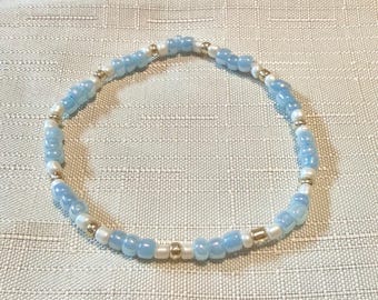 May include: A circular beaded bracelet featuring a repeating pattern of translucent light blue beads, opaque white beads, and small metallic gold-tone spacer beads. The beads are strung together in a continuous loop to form a simple, uniform design.