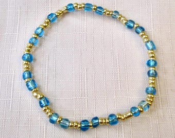 May include: A circular beaded bracelet featuring an alternating pattern of small, round, metallic gold-tone beads and slightly larger, translucent, light blue glass beads arranged in a continuous loop on a flexible cord.