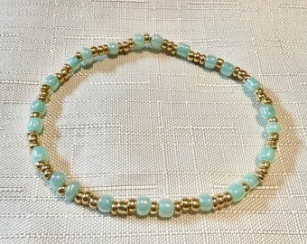 May include: A circular beaded bracelet featuring an alternating pattern of small, metallic gold-toned spherical beads and smooth, translucent light mint green beads. The beads are strung together in a continuous loop, creating a simple, uniform design.