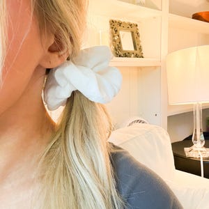 May include: A solid white fabric hair scrunchie with a gathered, voluminous texture. The accessory is secured around a ponytail of blonde hair, showcasing a soft, matte finish and a circular shape that creates a full, rounded appearance.