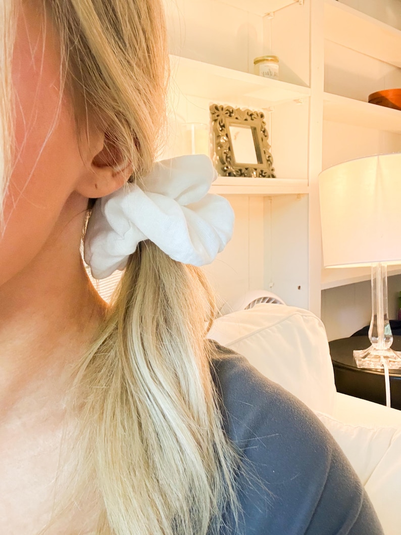 May include: A solid white fabric hair scrunchie with a gathered, voluminous texture. The accessory is secured around a ponytail of blonde hair, showcasing a soft, matte finish and a circular shape that creates a full, rounded appearance.