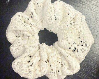 May include: A white hair scrunchie made from a delicate, open-weave lace fabric. The accessory features a gathered, circular design with a repeating geometric pattern throughout the material, creating a soft, textured, and voluminous appearance.