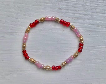 May include: A circular beaded bracelet featuring a repeating pattern of small, round, polished beads in alternating colors of vibrant red, soft pastel pink, and metallic gold. The beads are strung together on a flexible cord to form a simple loop.