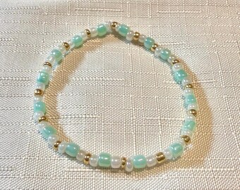May include: A circular beaded bracelet featuring an alternating pattern of small, round, translucent mint green beads, clear white beads, and tiny metallic gold-toned spacer beads arranged in a continuous, repeating sequence around the entire band.