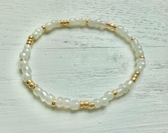 May include: A circular beaded bracelet featuring a repeating pattern of translucent white glass beads interspersed with small, metallic gold-toned spacer beads. The beads are strung together in a continuous loop, creating a simple and uniform design.