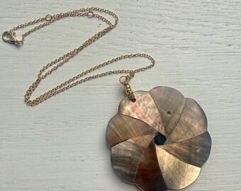 May include: A necklace featuring a thin gold-tone chain and a large, circular pendant composed of overlapping, iridescent brown and gray shell segments arranged in a spiral pattern, creating a flower-like shape with a small dark center.