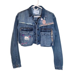 May include: A cropped blue denim jacket featuring a collar, long sleeves, and a button-up front. The jacket includes two chest pockets with zippers, decorative sequins, beaded fringe, and embroidered patches, including one that reads "HEY Y'ALL".