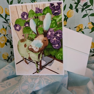 May include: A watercolour painting of a green and brown metal donkey sculpture standing in front of a wooden fence with purple morning glory flowers growing around it.