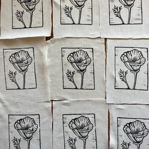 May include: Nine small, square fabric pieces with a black and white floral design. Each piece features a detailed illustration of a flower with a stem and leaves, framed within a black border. The fabric has a natural, off-white colour.