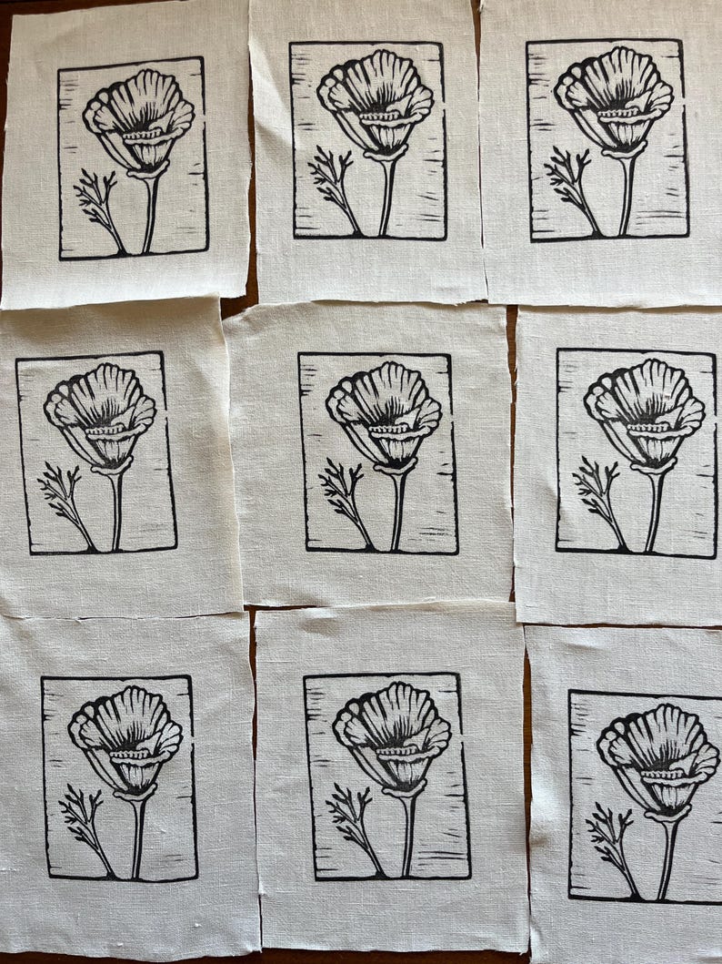 May include: Nine small, square fabric pieces with a black and white floral design. Each piece features a detailed illustration of a flower with a stem and leaves, framed within a black border. The fabric has a natural, off-white colour.