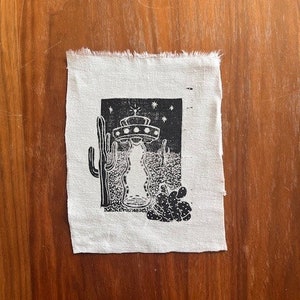 May include: A small, square, off-white fabric patch featuring a black and white linocut print. The print depicts a UFO hovering over a desert landscape with cacti and stars. The fabric has a raw, unfinished edge.