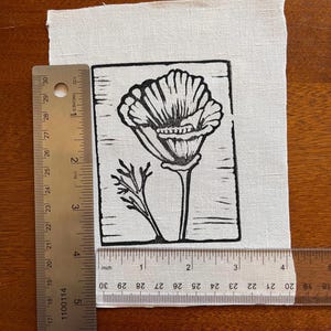 May include: A black and white linocut print of a poppy flower on a white fabric. The print is approximately 10 cm wide and 13 cm tall. The poppy has detailed petals and a stem with leaves. The fabric is rectangular and has a slightly textured surface.