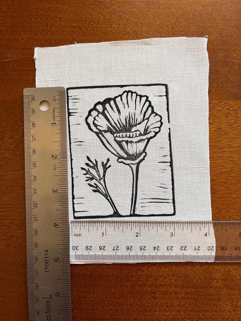 May include: A black and white linocut print of a poppy flower on a white fabric. The print is approximately 10 cm wide and 13 cm tall. The poppy has detailed petals and a stem with leaves. The fabric is rectangular and has a slightly textured surface.
