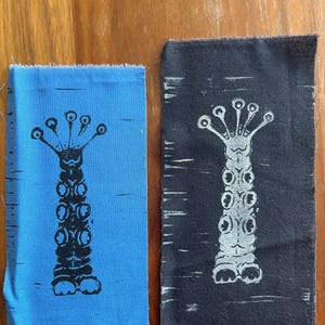 May include: Two fabric patches with a black and white print of a stylized creature with multiple eyes and a segmented body. The creature is printed on a blue patch and a black patch.