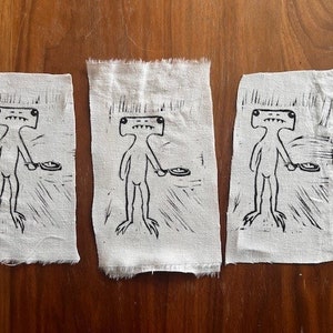 Pu&ograve; includere: Tre toppe di tessuto bianco con illustrazioni a inchiostro nero di una figura umanoide con una testa grande e un piatto in mano. La figura ha un corpo lungo e sottile e occhi grandi e rotondi.