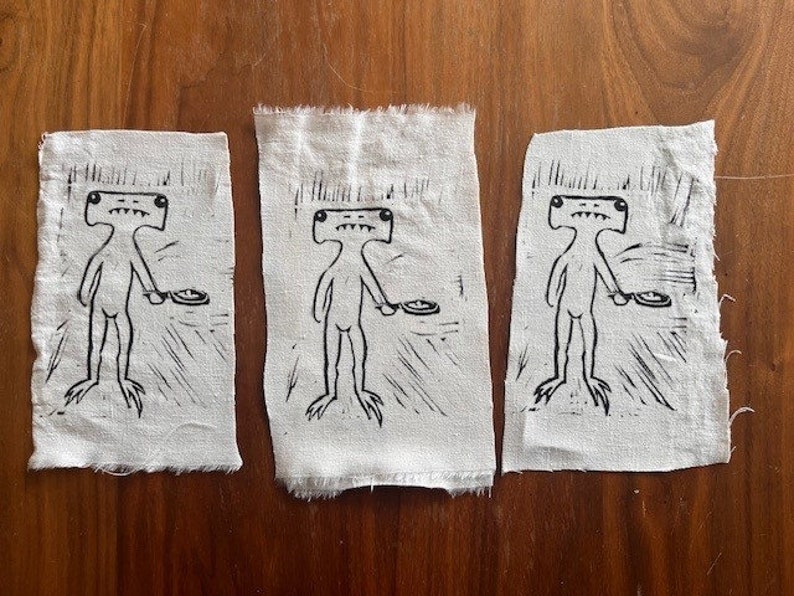 Pu&ograve; includere: Tre toppe di tessuto bianco con illustrazioni a inchiostro nero di una figura umanoide con una testa grande e un piatto in mano. La figura ha un corpo lungo e sottile e occhi grandi e rotondi.