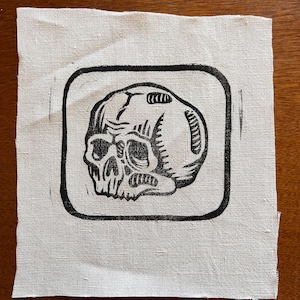 May include: A black and white linocut print of a skull within a square border. The skull is detailed with visible eye sockets, teeth, and cranial features. The print is on a square piece of off-white fabric.