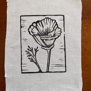 May include: A black and white linocut print of a poppy flower and foliage on off-white fabric. The poppy is in full bloom, with detailed petals and a long stem. The print is framed by a rectangular border.