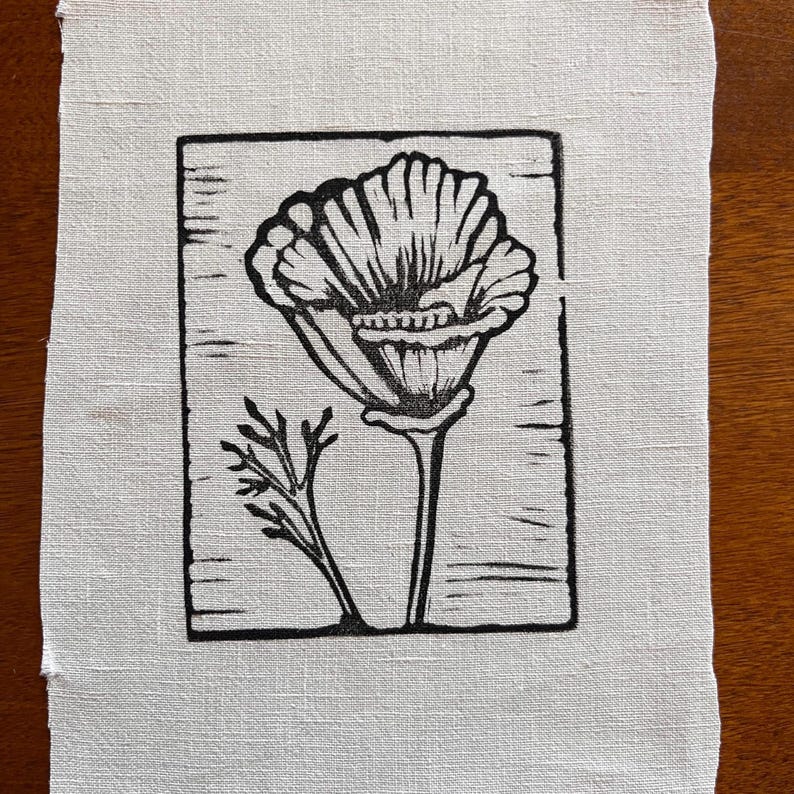 May include: A black and white linocut print of a poppy flower and foliage on off-white fabric. The poppy is in full bloom, with detailed petals and a long stem. The print is framed by a rectangular border.