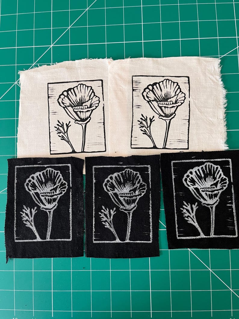 May include: Five fabric patches featuring a poppy flower design. Two are on cream-coloured fabric, and three are on black fabric. The poppy design is in black and white, with a detailed flower and stem.
