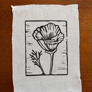May include: A black and white linocut print of a flower on a square piece of off-white fabric. The flower is in full bloom, with detailed petals and a stem. A smaller sprig of leaves is to the left of the flower. The fabric has a raw edge.