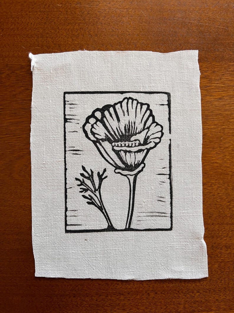 May include: A black and white linocut print of a flower on a square piece of off-white fabric. The flower is in full bloom, with detailed petals and a stem. A smaller sprig of leaves is to the left of the flower. The fabric has a raw edge.