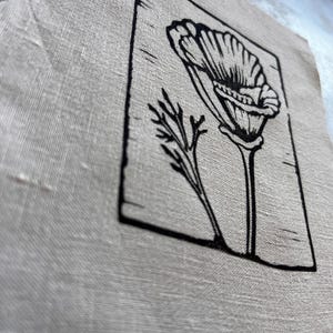May include: A piece of beige linen fabric with a black floral linocut print. The print shows a detailed illustration of a flower and a sprig of leaves within a rectangular frame. The fabric has a slightly frayed edge.