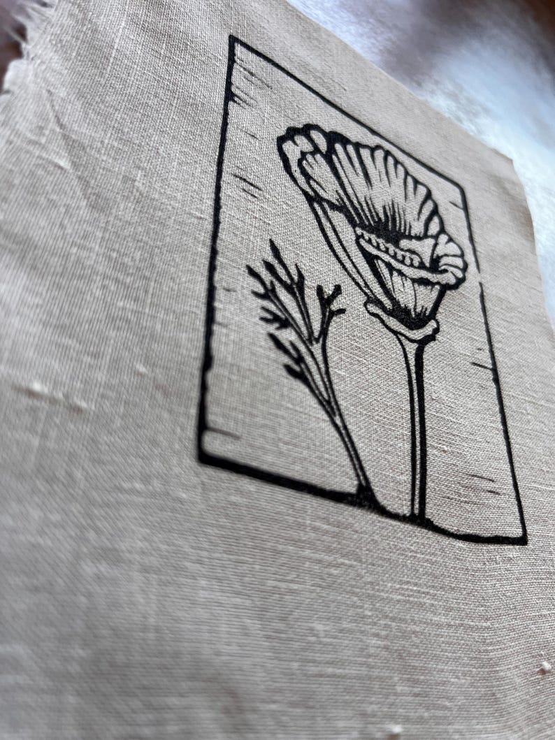 May include: A piece of beige linen fabric with a black floral linocut print. The print shows a detailed illustration of a flower and a sprig of leaves within a rectangular frame. The fabric has a slightly frayed edge.