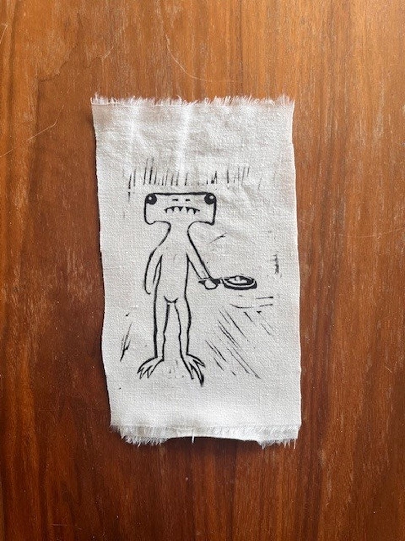 Pu&ograve; includere: Un'illustrazione in bianco e nero di una creatura che tiene una padella. La creatura ha una testa grande con due occhi grandi e una bocca larga. La creatura sta in piedi su due gambe e ha due braccia. La creatura tiene una padella in una mano. L'illustrazione &egrave; su uno sfondo di tessuto bianco.